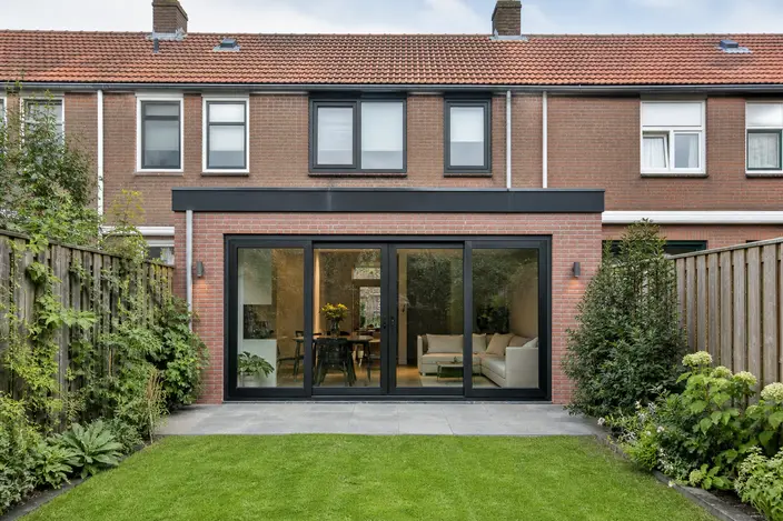 AI-beeld: een nieuwe aanbouw van baksteen met een plat dak en grote glazen deuren aan de achterkant van een Nederlands rijtjeshuis, met een tuin ervoor